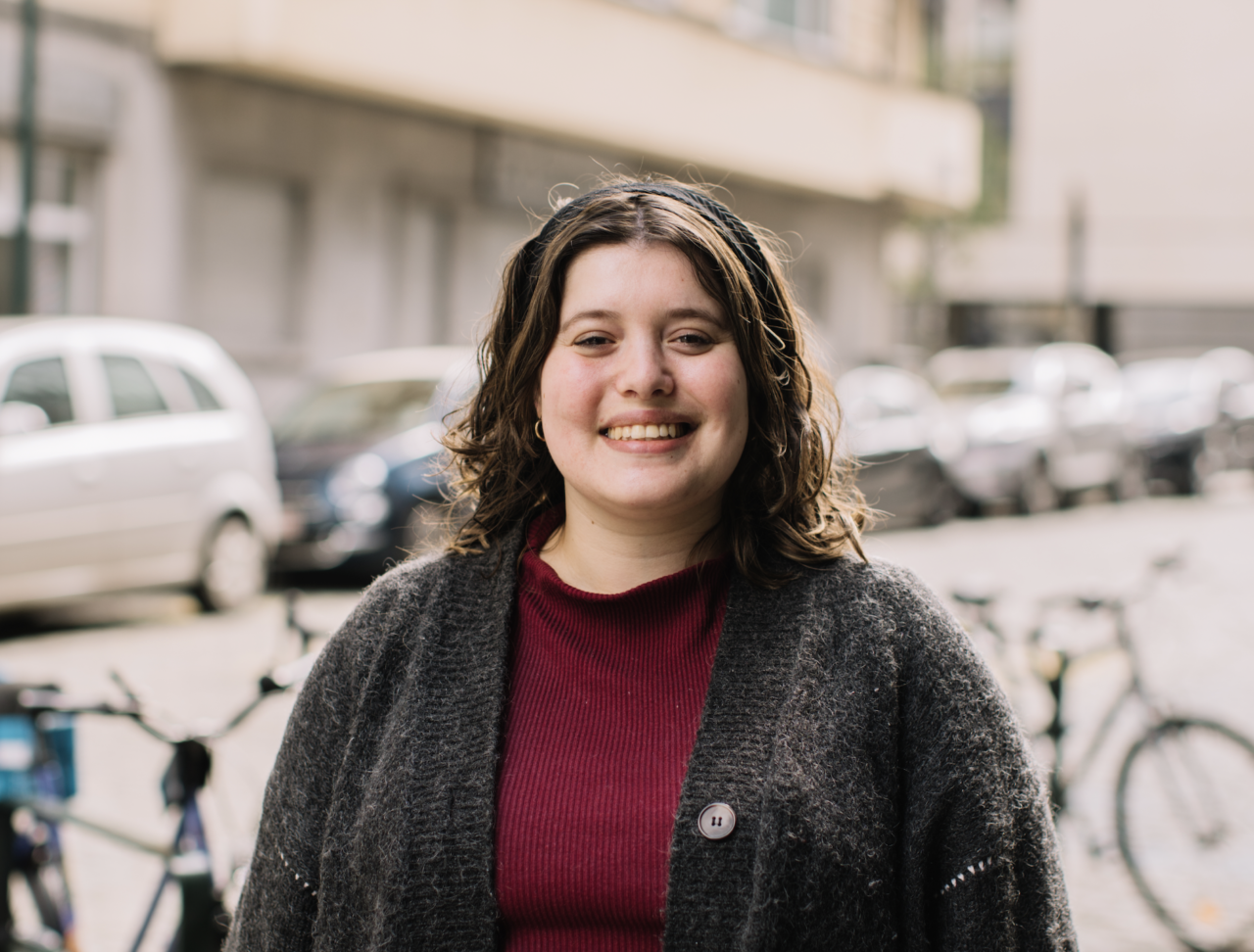 Woman, smiling in front of the camera, in the streets of Brussels. There are bikes and cars behind. She is wearing a red t-shirt and grey sweatshirt