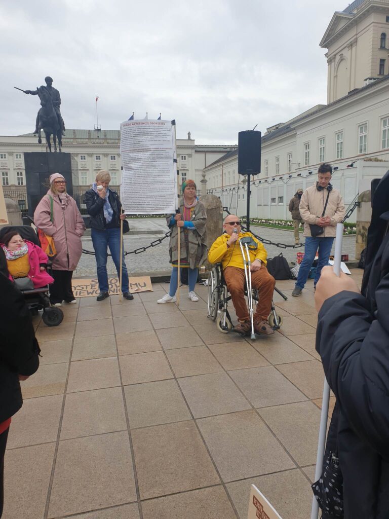 People are in a square, someone is speaking in a microphone and people are standing on the side.