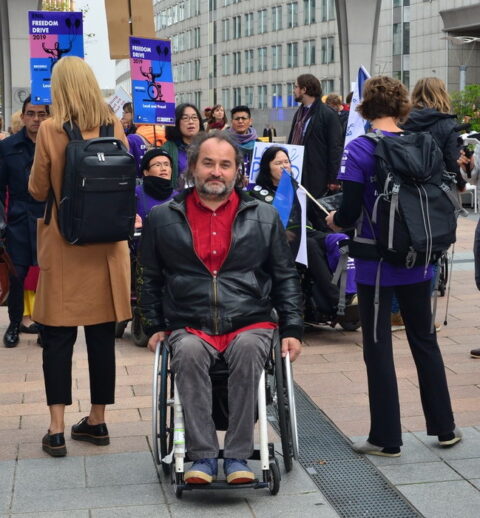 A man ina wheelchair, looking at the camera, in front of a crowd holding banners.