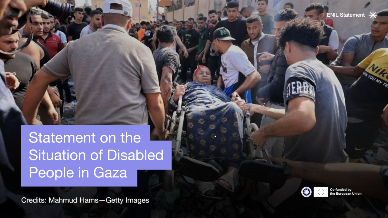 Statement on the situation of Disabled People in Gaza. photo of a woman in a wheelchair being carried through the crowd and rubble.