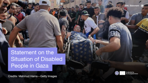 Statement on the situation of Disabled People in Gaza. photo of a woman in a wheelchair being carried through the crowd and rubble.