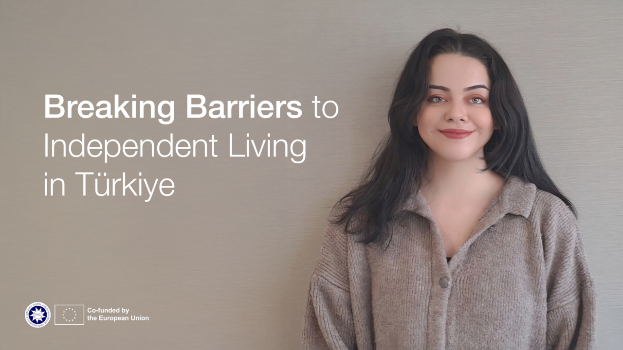 "Breaking Barriers to Independent Living in Turkiye". I am a woman in my late 20s with light skin and straight, shoulder-length black hair. I am wearing a textured, light brown button-up sweater with a relaxed fit. My expression is warm and approachable, with a slight smile. I am not wearing any visible accessories, such as glasses or jewelry, and I am not using any mobility aids. The background is plain and neutral, keeping the focus on me.