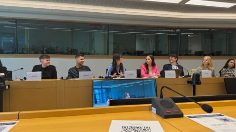 Panel discussion at the European Parliament with several speakers seated behind a long desk, each with a microphone and nameplate. Participants are engaged in conversation, with one person speaking while others listen and smile. Interpretation booths are visible in the background, and a screen in the foreground shows remote participants, indicating a hybrid event.