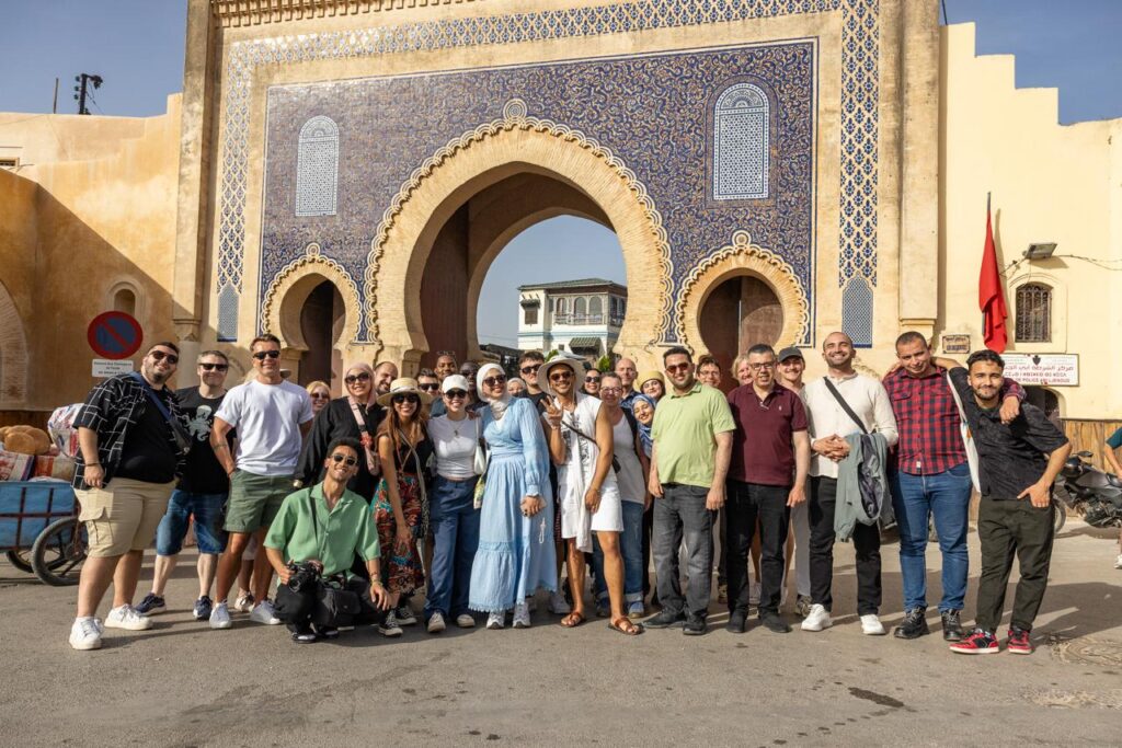 Alt text: A large group of people posing for a photo in front of an ornate historic gate with blue and gold tilework and large arched entrances. The group is standing outdoors in bright sunlight, smiling at the camera. A red flag, street signs, parked vehicles, and surrounding beige buildings are visible in the background.