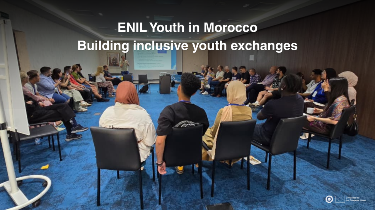Alt text: A group of people sit in a circle during a training session in Morocco. A presenter stands at the front of the room next to a projected presentation. White text at the top of the image reads: “ENIL Youth in Morocco. Building inclusive youth exchanges.”