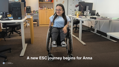 Alt text: Anna sits in a wheelchair in an ENIL office, smiling towards the camera. She is wearing a light blue top, dark trousers and black trainers. White text over the lower part of the image reads: “A new ESC journey begins for Anna.” The ENIL and EU co-funded logos appear in the bottom left corner.