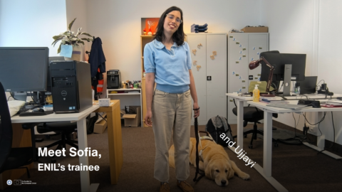 Sofia stands in an office, smiling at the camera, with her guide dog Ujjayi lying on the floor beside her. White text on the lower left reads: “Meet Sofia, ENIL’s new trainee.” Another white text label near the dog reads: “and Ujjayi.”