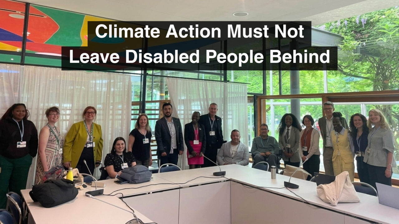 Here’s a clear alt text version: A group of conference participants pose together in a meeting room at a climate conference. Some participants are seated and some are standing, including wheelchair users. Large white text on black background reads: “Climate Action Must Not Leave Disabled People Behind.”